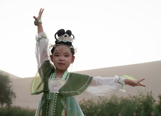 A young girl is dressed in traditional attire, wearing a green and white flowing dress with decorative patterns. Her hair is adorned with intricate accessories, including a delicate, jeweled headpiece. She poses gracefully with one arm raised, exuding a sense of elegance and poise. The background is blurred with soft, natural tones, suggesting an outdoor setting.