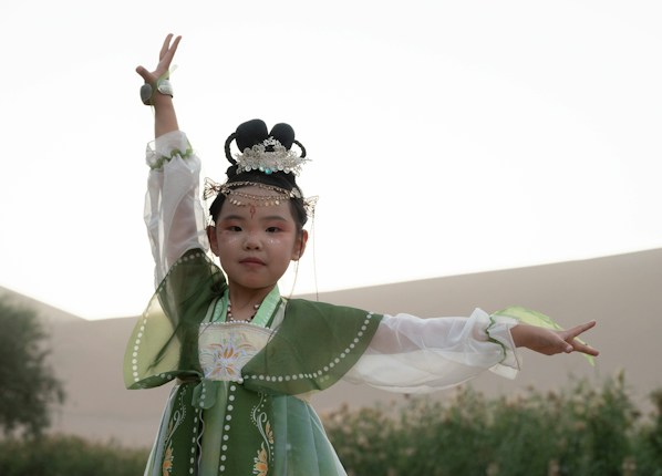 A young girl is dressed in traditional attire, wearing a green and white flowing dress with decorative patterns. Her hair is adorned with intricate accessories, including a delicate, jeweled headpiece. She poses gracefully with one arm raised, exuding a sense of elegance and poise. The background is blurred with soft, natural tones, suggesting an outdoor setting.