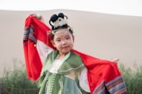 A child dressed in traditional clothing stands with arms raised, holding a red cloak. The attire is ornate, with intricate patterns and accessories, including hair ornaments and face paint. The background features a neutral sand dune and foliage.