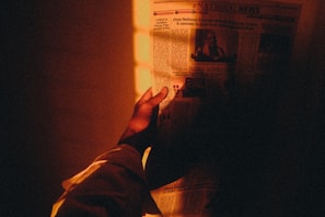 A hand holds a newspaper under warm, dim lighting. The newspaper text and images are partially visible, creating a cozy, intimate atmosphere with light casting an amber glow.