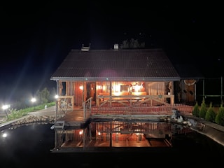 A wooden cabin is illuminated warmly at night, with lights reflecting on a nearby water body. The cabin features a shingled roof and a wooden deck area surrounded by a small fence. There are ornamental plants lining the pathway to the cabin.