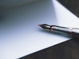 A close-up of a fountain pen with a silver and black nib resting on a blank sheet of paper. The pen's metallic components have a polished finish, reflecting light subtly. The scene is captured on a smooth, soft-focus background, creating a calm and gentle atmosphere.