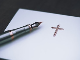 A close-up of a fountain pen placed next to an intricately designed cross on a piece of paper. The pen has a metallic body with copper-colored accents.