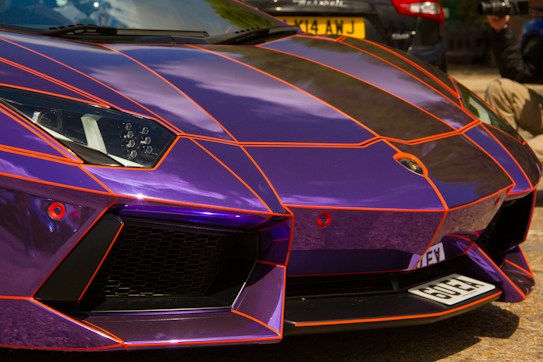 The image captures a close-up view of the front of a luxurious sports car featuring a glossy purple paint job accentuated with bright orange stripes in a geometric pattern. The car's sleek design, sharp angles, and intricate headlight details are prominent. A portion of a photographer or videographer is visible in the background, emphasizing the car's high-profile appeal.