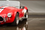 A classic red convertible car with white racing stripes is parked in a garage setting. The bodywork is shiny with water droplets, indicating recent rainfall. The car has prominent chrome bumpers and round headlights, with detailed tire treads visible on the front wheels.
