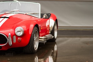 A classic red convertible car with white racing stripes is parked in a garage setting. The bodywork is shiny with water droplets, indicating recent rainfall. The car has prominent chrome bumpers and round headlights, with detailed tire treads visible on the front wheels.