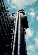 A modern architectural structure with prominent metallic pipes extending upward against the backdrop of a partly cloudy sky. The pipes are organized in parallel and appear to be part of an industrial or futuristic building exterior.