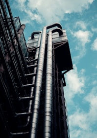 A modern architectural structure with prominent metallic pipes extending upward against the backdrop of a partly cloudy sky. The pipes are organized in parallel and appear to be part of an industrial or futuristic building exterior.