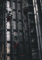 A series of modern, metallic elevators on the exterior of a building, with visible metal beams and glass components. Red lights are illuminated on several elevator cars, punctuating the industrial aesthetic. The structure features a sleek, dark design with reflections hinting at the surrounding architecture.