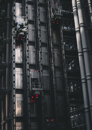 A series of modern, metallic elevators on the exterior of a building, with visible metal beams and glass components. Red lights are illuminated on several elevator cars, punctuating the industrial aesthetic. The structure features a sleek, dark design with reflections hinting at the surrounding architecture.