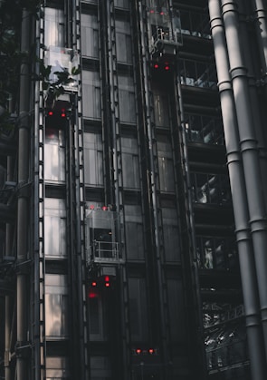 A series of modern, metallic elevators on the exterior of a building, with visible metal beams and glass components. Red lights are illuminated on several elevator cars, punctuating the industrial aesthetic. The structure features a sleek, dark design with reflections hinting at the surrounding architecture.