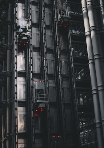 A series of modern, metallic elevators on the exterior of a building, with visible metal beams and glass components. Red lights are illuminated on several elevator cars, punctuating the industrial aesthetic. The structure features a sleek, dark design with reflections hinting at the surrounding architecture.