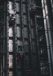 A series of modern, metallic elevators on the exterior of a building, with visible metal beams and glass components. Red lights are illuminated on several elevator cars, punctuating the industrial aesthetic. The structure features a sleek, dark design with reflections hinting at the surrounding architecture.