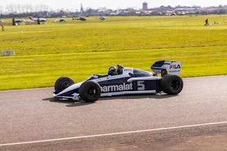 A vintage Formula 1 racing car is driving on a track. The sleek black and white design features prominent sponsor logos like Parmalat and Michelin. The background shows a field with various small planes and a distant townscape under a clear blue sky. The car appears to be moving at speed, creating a sense of dynamic motion.