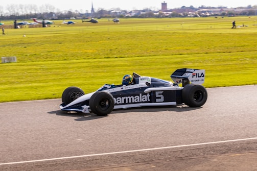 A vintage Formula 1 racing car is driving on a track. The sleek black and white design features prominent sponsor logos like Parmalat and Michelin. The background shows a field with various small planes and a distant townscape under a clear blue sky. The car appears to be moving at speed, creating a sense of dynamic motion.