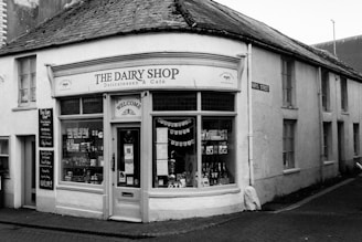 A quaint corner shop with a rustic exterior, displaying signage for 'The Dairy Shop' and 'Delicatessen & Café'. The shop windows showcase a variety of products, including bottles and packages. The building features white walls and a slate roof, situated on a narrow cobblestone street. The façade is decorated with small welcome signs and strings of bunting.