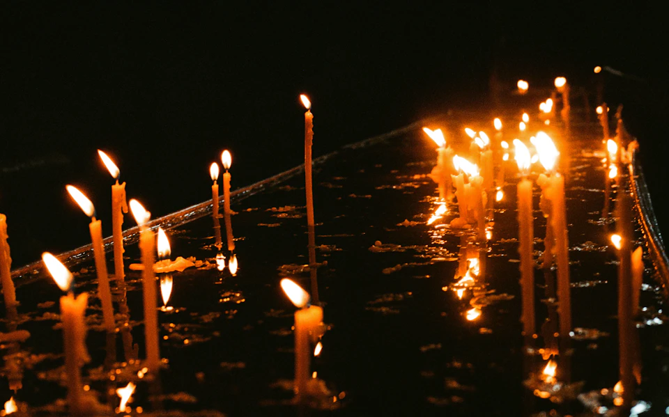 The image depicts a group of candles, each with its own unique shape and size, arranged on a black surface. The candles are lit from below, casting a warm glow that illuminates their surroundings. Some of the candles have been partially extinguished, adding an element of depth to the scene. The background is dark, allowing the candles to stand out prominently against it.