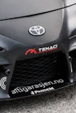 The image features the front part of a car with a focus on the bumper and grill area. Prominently displayed is the Toyota logo in the center of the car's hood. Nearby, there are several decals, including the words 'TENACI Parts for Racecars' in white with a red gear graphic, and 'altigarasjen.no' along with the 'Pandem' logo.