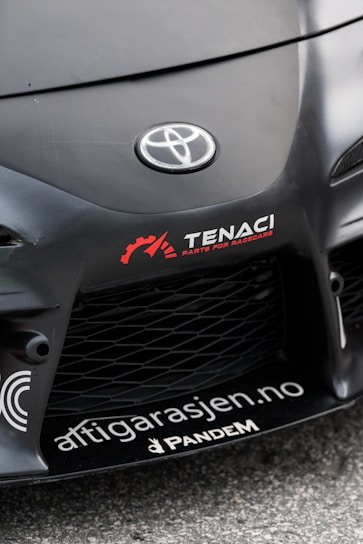 The image features the front part of a car with a focus on the bumper and grill area. Prominently displayed is the Toyota logo in the center of the car's hood. Nearby, there are several decals, including the words 'TENACI Parts for Racecars' in white with a red gear graphic, and 'altigarasjen.no' along with the 'Pandem' logo.