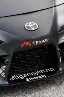 The image features the front part of a car with a focus on the bumper and grill area. Prominently displayed is the Toyota logo in the center of the car's hood. Nearby, there are several decals, including the words 'TENACI Parts for Racecars' in white with a red gear graphic, and 'altigarasjen.no' along with the 'Pandem' logo.