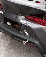 Close-up of the rear section of a race car featuring a prominently displayed logo and branding for a motorsport company. The car's exterior is matte black with a visible tow hook and some custom aerodynamic modifications.