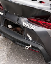 Close-up of the rear section of a race car featuring a prominently displayed logo and branding for a motorsport company. The car's exterior is matte black with a visible tow hook and some custom aerodynamic modifications.