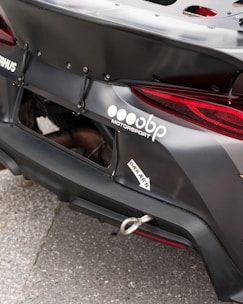 Close-up of the rear section of a race car featuring a prominently displayed logo and branding for a motorsport company. The car's exterior is matte black with a visible tow hook and some custom aerodynamic modifications.