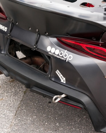Close-up of the rear section of a race car featuring a prominently displayed logo and branding for a motorsport company. The car's exterior is matte black with a visible tow hook and some custom aerodynamic modifications.