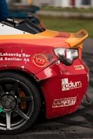 A close-up of a damaged sports car with a vibrant red and orange rear section. The car features several logos and text, including 'VVS Fagmann' and 'Bergen Gokart'. The vehicle has a visible dent and scratches near the wheel, indicating it has been involved in some rough activity. The wheel is gray with a five-spoke design. The scene is likely at a racing event, suggested by the setting and condition of the car.