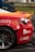 A close-up of a damaged sports car with a vibrant red and orange rear section. The car features several logos and text, including 'VVS Fagmann' and 'Bergen Gokart'. The vehicle has a visible dent and scratches near the wheel, indicating it has been involved in some rough activity. The wheel is gray with a five-spoke design. The scene is likely at a racing event, suggested by the setting and condition of the car.