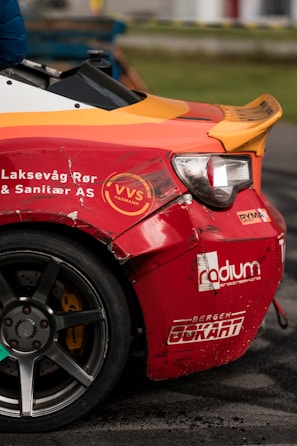 A close-up of a damaged sports car with a vibrant red and orange rear section. The car features several logos and text, including 'VVS Fagmann' and 'Bergen Gokart'. The vehicle has a visible dent and scratches near the wheel, indicating it has been involved in some rough activity. The wheel is gray with a five-spoke design. The scene is likely at a racing event, suggested by the setting and condition of the car.