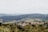 A picturesque landscape of rolling hills with lush green vineyards and scattered trees. A quaint village with red-roofed houses nestles among the greenery, evoking the charm of the Italian countryside. The background features layered hills receding into the distance under a hazy sky.