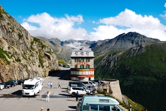 A vintage hotel building is situated on a mountain road with a backdrop of breathtaking alpine scenery. The area is surrounded by rocky cliffs and lush green hills under a bright blue sky. Cars and a bus are parked in the foreground, and a few people are walking around, enjoying the stunning view.
