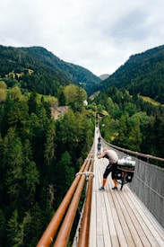 A long suspension bridge stretches over a lush, forested valley with tall trees and rolling hills in the background. A person is seen on the bridge with a bicycle, pausing to take a photo. The surrounding landscape is vibrant with greenery, and the sky appears overcast.