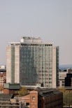 A tall, modern office building with a rectangular structure and a series of glass windows, located in an urban environment. In the foreground, there are older brick buildings, while the background features a clear sky and a distant body of water.