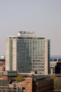A tall, modern office building with a rectangular structure and a series of glass windows, located in an urban environment. In the foreground, there are older brick buildings, while the background features a clear sky and a distant body of water.