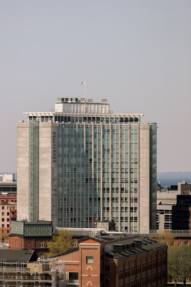 A tall, modern office building with a rectangular structure and a series of glass windows, located in an urban environment. In the foreground, there are older brick buildings, while the background features a clear sky and a distant body of water.