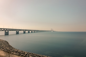 A long bridge stretches across calm water, connecting two distant points. The sky is clear with a hint of mist, and the horizon is barely visible. The bridge is supported by a series of pillars and extends from a rocky shoreline.