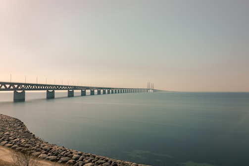 A long bridge stretches across calm water, connecting two distant points. The sky is clear with a hint of mist, and the horizon is barely visible. The bridge is supported by a series of pillars and extends from a rocky shoreline.