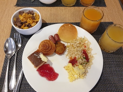 A variety of breakfast foods arranged on a white plate, including scrambled eggs, noodles with sauce, a small pancake, a sausage, a pastry spiral, and a slice of cake with red jelly. A bowl of cereal beside the plate. Several glasses of orange juice are also on the table, and silverware is placed next to the plate on a woven mat.