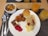 A variety of breakfast foods arranged on a white plate, including scrambled eggs, noodles with sauce, a small pancake, a sausage, a pastry spiral, and a slice of cake with red jelly. A bowl of cereal beside the plate. Several glasses of orange juice are also on the table, and silverware is placed next to the plate on a woven mat.