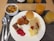 A variety of breakfast foods arranged on a white plate, including scrambled eggs, noodles with sauce, a small pancake, a sausage, a pastry spiral, and a slice of cake with red jelly. A bowl of cereal beside the plate. Several glasses of orange juice are also on the table, and silverware is placed next to the plate on a woven mat.