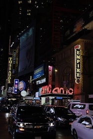 A bustling urban street scene at night, featuring bright neon lights and signs from various businesses. Cars are visible on the street, and there are advertisements and marquees for entertainment venues, including a large AMC theater sign.