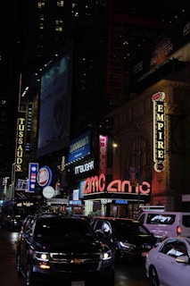 A bustling urban street scene at night, featuring bright neon lights and signs from various businesses. Cars are visible on the street, and there are advertisements and marquees for entertainment venues, including a large AMC theater sign.