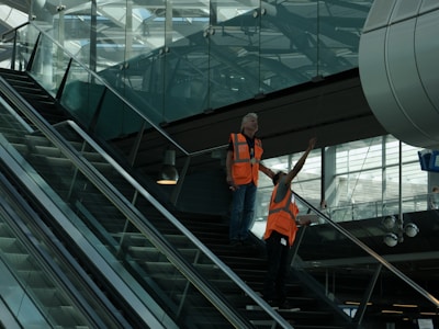 Two individuals wearing bright orange safety vests are standing on an escalator inside a modern building with large glass windows. One person is pointing upwards, and the environment appears to be industrial or related to transportation.