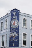 A tall, painted blue sign on the side of a light-colored building features ornate text and design elements. The sign advertises a distillery, prominently featuring the words 'Home of Portobello Road London Dry Gin' and 'Proud Purveyors of London Spirit.' Decorative elements include a red lion emblem and stylized fonts.