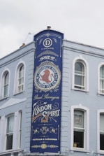 A tall, painted blue sign on the side of a light-colored building features ornate text and design elements. The sign advertises a distillery, prominently featuring the words 'Home of Portobello Road London Dry Gin' and 'Proud Purveyors of London Spirit.' Decorative elements include a red lion emblem and stylized fonts.