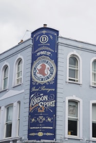 A tall, painted blue sign on the side of a light-colored building features ornate text and design elements. The sign advertises a distillery, prominently featuring the words 'Home of Portobello Road London Dry Gin' and 'Proud Purveyors of London Spirit.' Decorative elements include a red lion emblem and stylized fonts.
