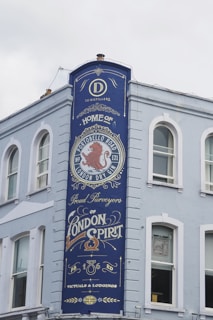A tall, painted blue sign on the side of a light-colored building features ornate text and design elements. The sign advertises a distillery, prominently featuring the words 'Home of Portobello Road London Dry Gin' and 'Proud Purveyors of London Spirit.' Decorative elements include a red lion emblem and stylized fonts.