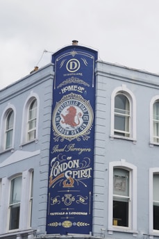 A tall, painted blue sign on the side of a light-colored building features ornate text and design elements. The sign advertises a distillery, prominently featuring the words 'Home of Portobello Road London Dry Gin' and 'Proud Purveyors of London Spirit.' Decorative elements include a red lion emblem and stylized fonts.